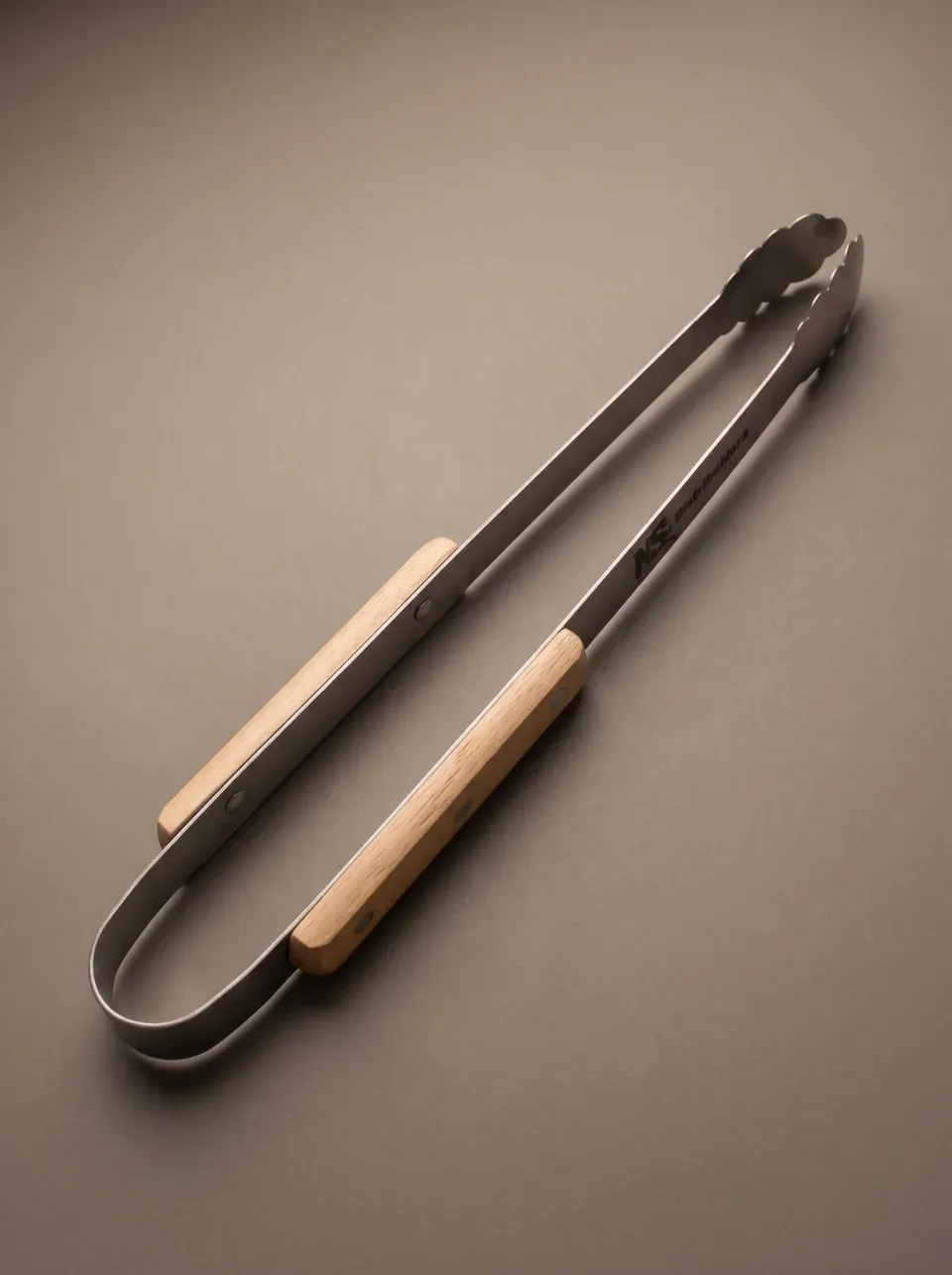 Pegador de churrasco de haste longa em aço inox, com dois cabos de madeira clara e pinça serrilhada, gravado a laser na haste.